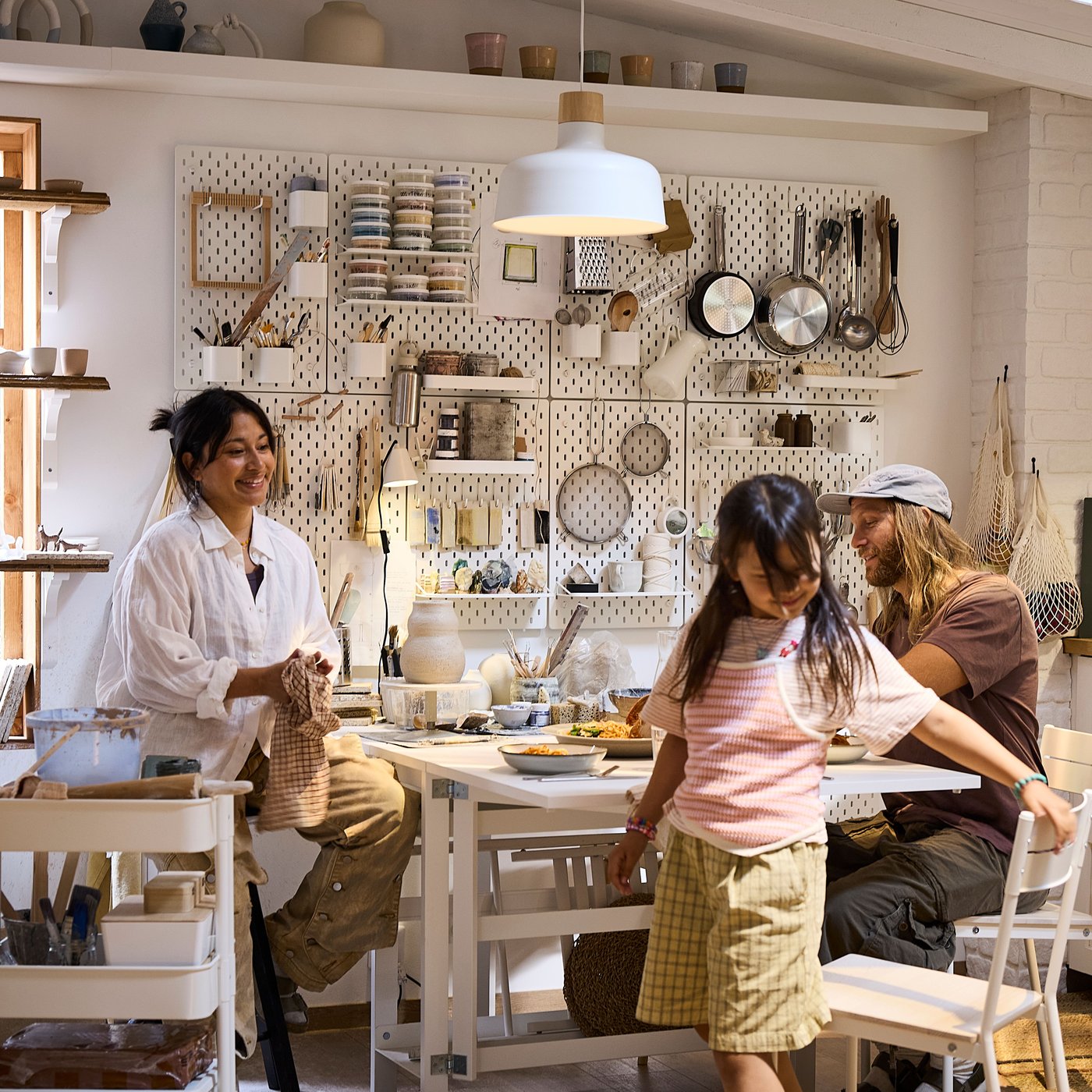 A family of three sits down to eat dinner together in a kitchen that’s also a hobby pottery studio with lots of wall storage.