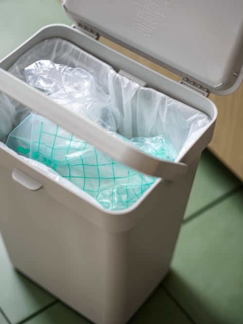 A light-grey HÅLLBAR waste-sorting bin standing on a green-tiled floor, holding discarded bits of plastic.