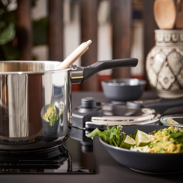 Oală de inox cu lingură de lemn pe aragaz; în prim-plan, bol cu mâncare decorată cu verdeață și felii de lămâie. În fundal se văd vase și obiecte decorative.