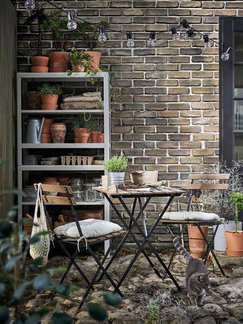Zonă exterioară cu mobilier cu două scaune și o masă, o unitate de rafturi, lanț de iluminat cu LED-uri, ghivece, plante în ghivece, o pisică.