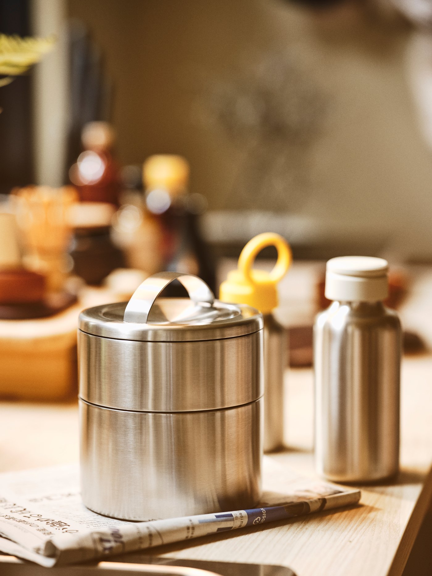 A wooden surface with a newspaper under a FÖRSKAFFA insulated tiffin box and an ENKELSPÅRIG stainless steel water bottle.