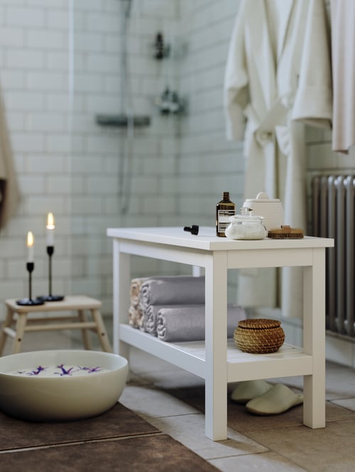 A white HEMNES bench in a white tiled bathroom with rolled-up towels and a basket on the lower shelf and containers on top.