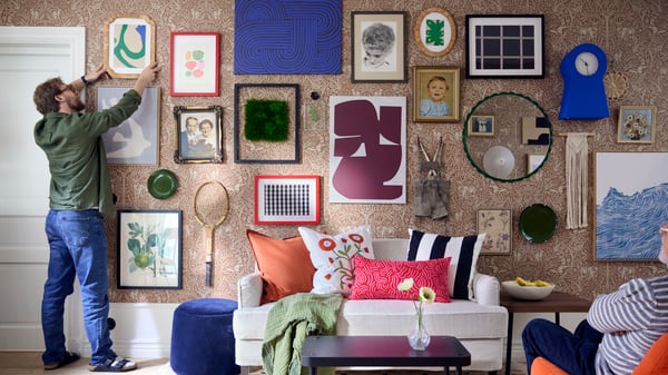A man hangs a picture frame on a wall decorated with several art pieces, such as posters, picture frames, and mirrors, in the living room.