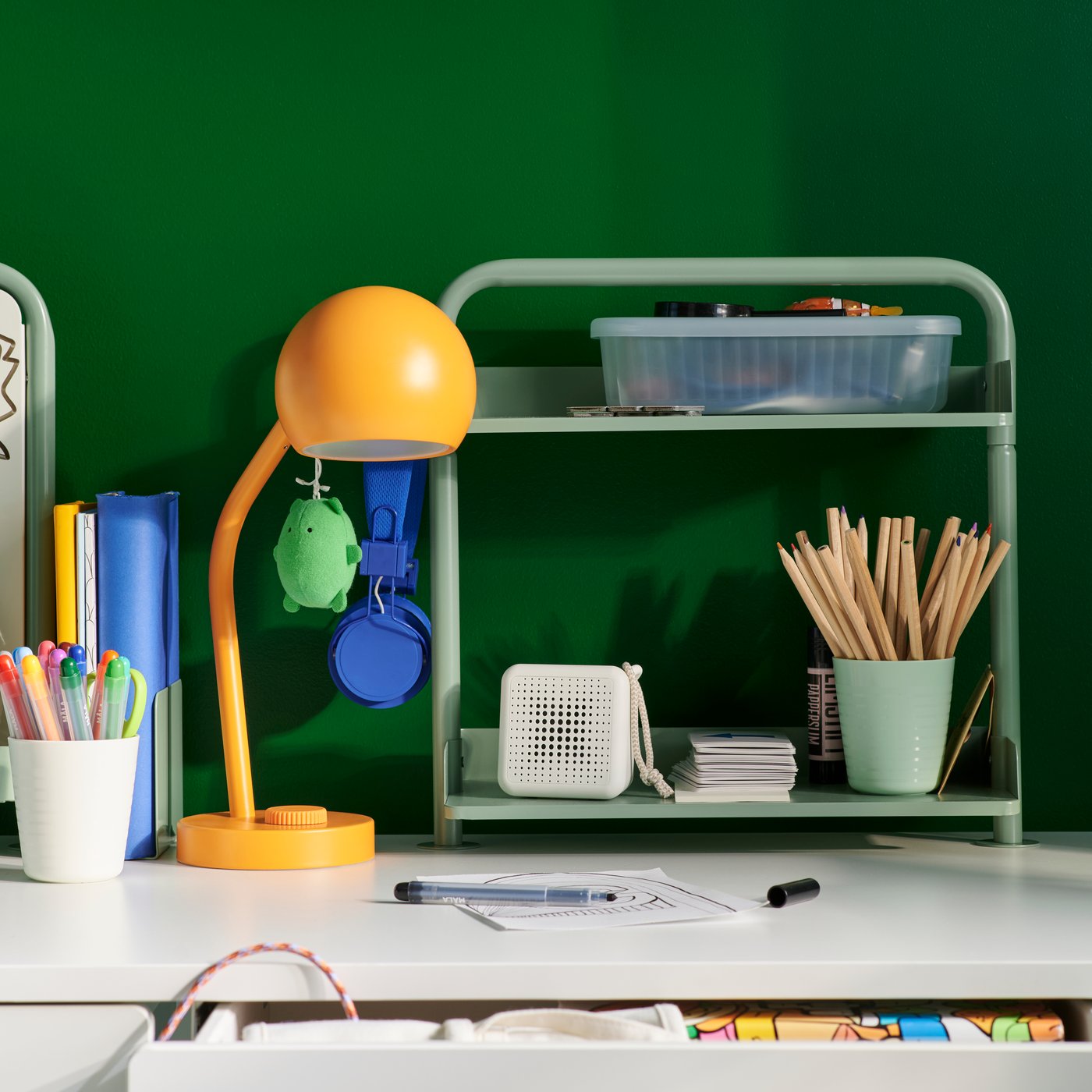 A RELATERA desk top shelf in green with two tiers holding stationery and a storage box, on a white desk against a green wall.