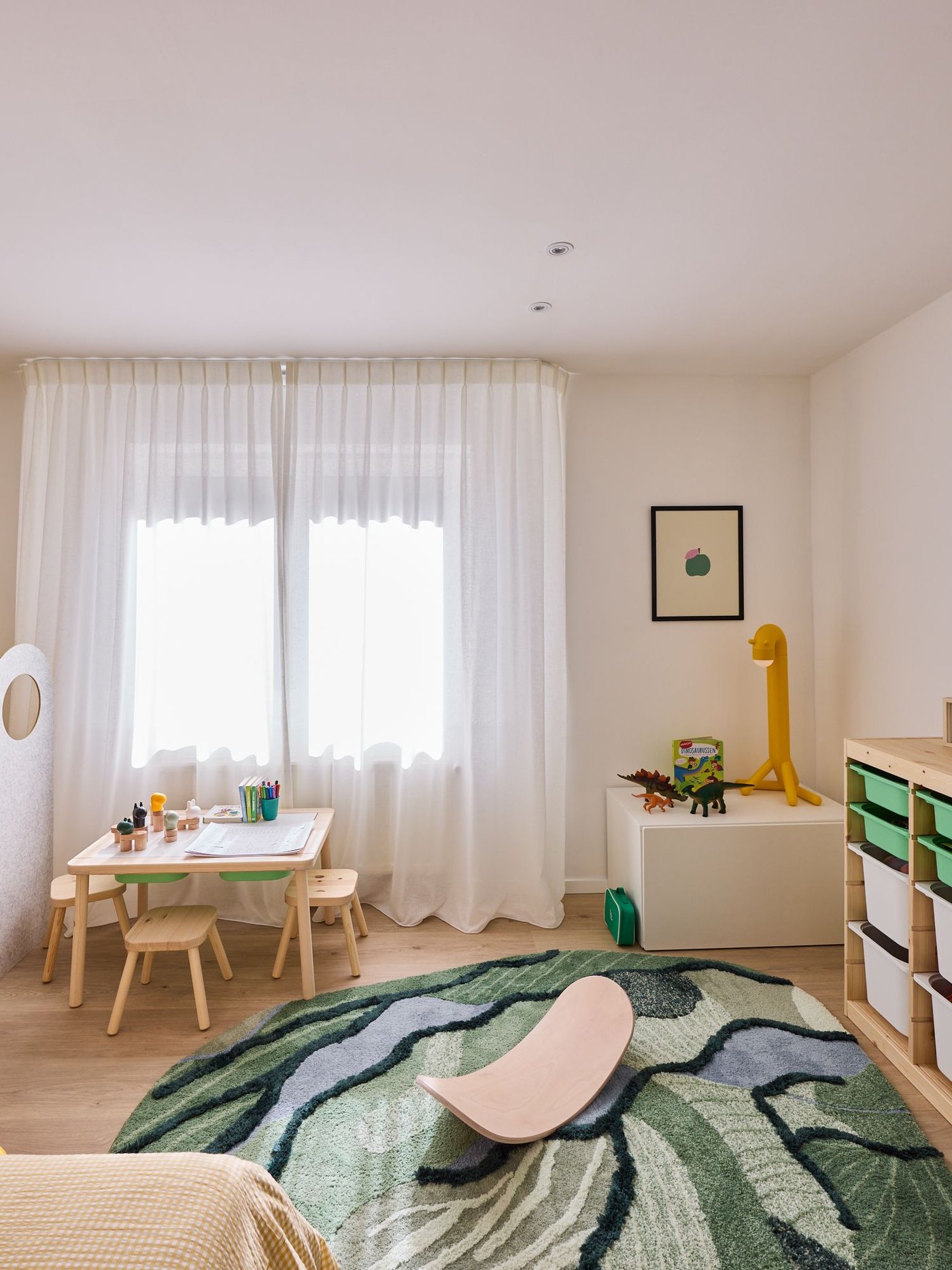 Lichte kinderkamer met speeltafel, opbergkasten, groen tapijt en speelgoed, zacht daglicht door gordijnen.