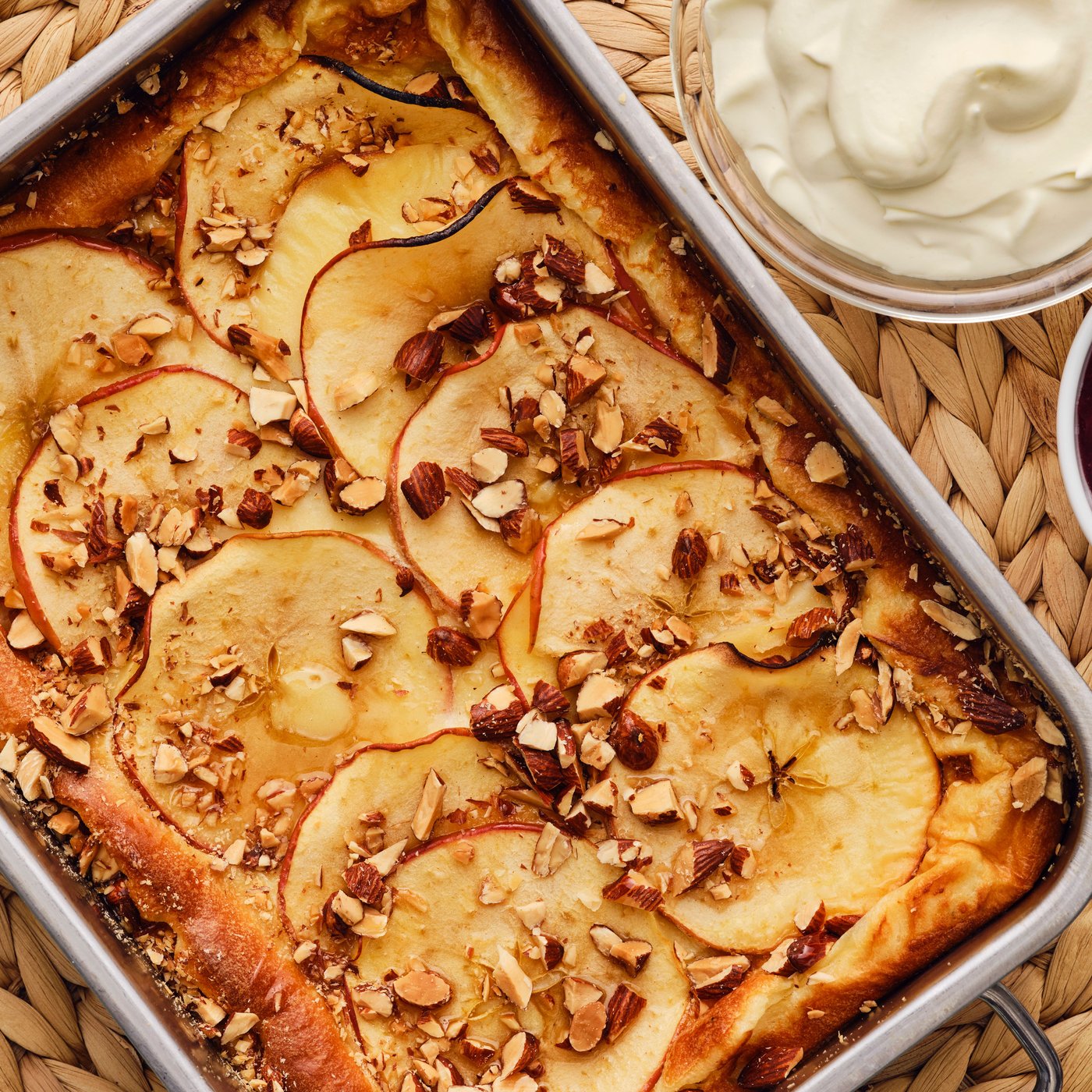 A baked golden-brown oven pancake with apples and almonds is served in a metal baking tray next to a bowl with whipped cream.