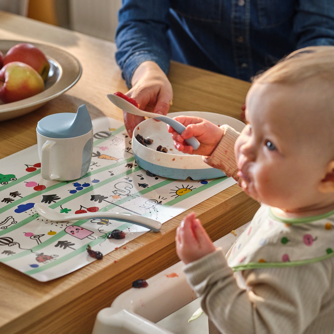 A child holds a BÖRJA feeding spoon sits in an ANTILOP armchair that’s at a dining table with a BÖRJA plate and beaker on.