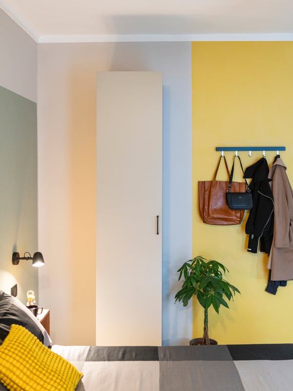 Una parete di una camera da letto con un mobile alto e attaccapanni TJUSIG a cui sono appesi degli abiti e due borse. Il letto con lenzuola, con fantasie geometriche e un cuscino giallo, è illuminato da una lampada parete SKURUP.