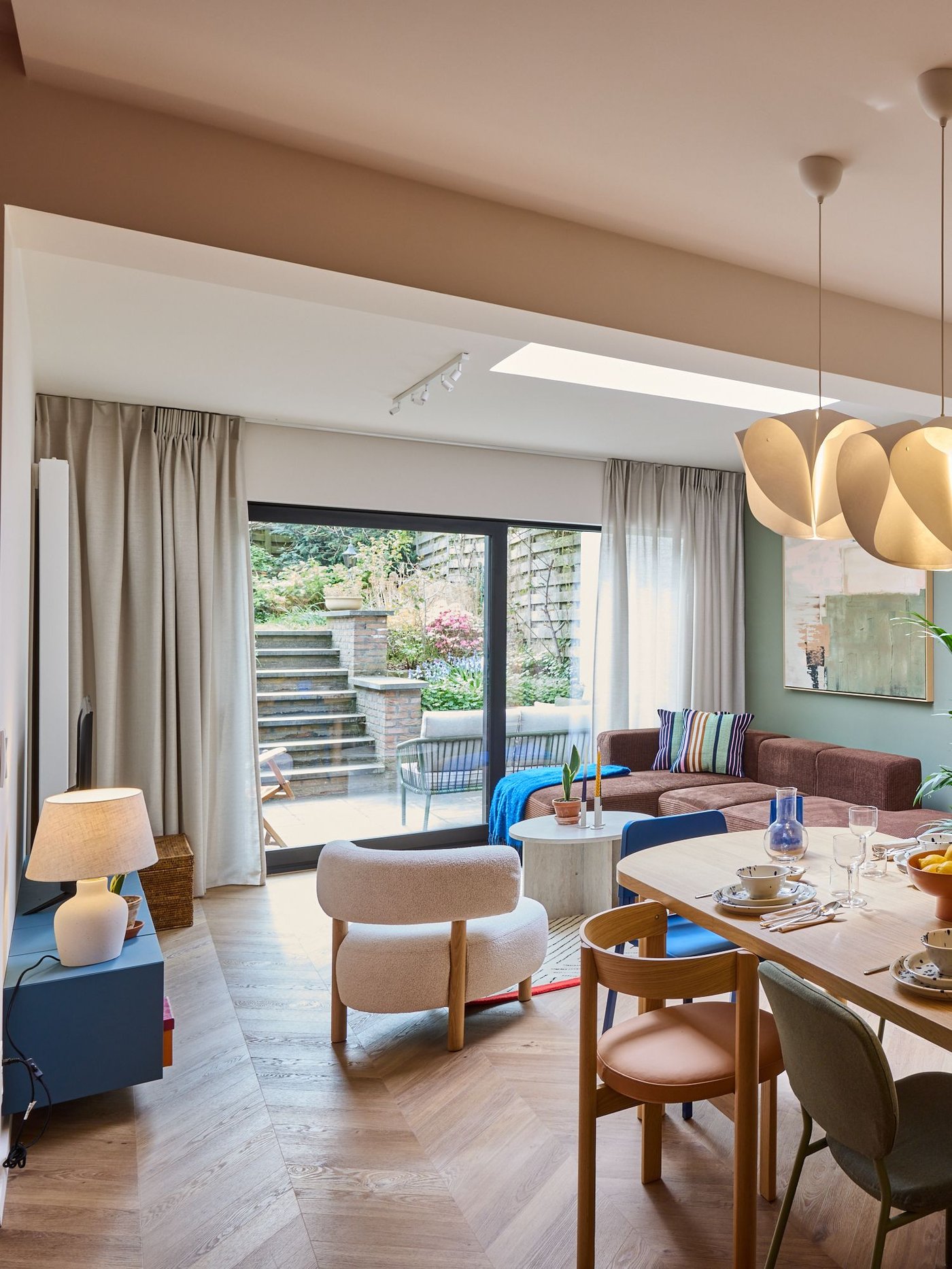 Lichte woonkamer met eettafel, stoelen en zetel, grote schuiframen naar tuin, houten vloer en warme verlichting in modern interieur.