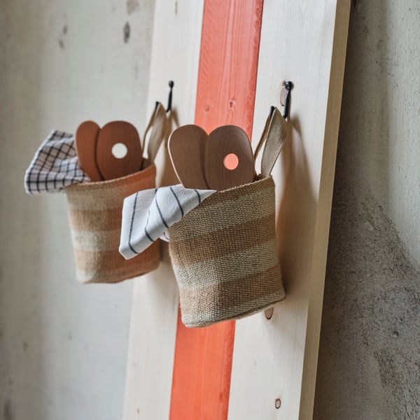 Două coșuri de depozitare MÄVINN din iută, umplute cu ustensile din lemn și șervete de masă, sunt agățate în cârlige pe un perete.
