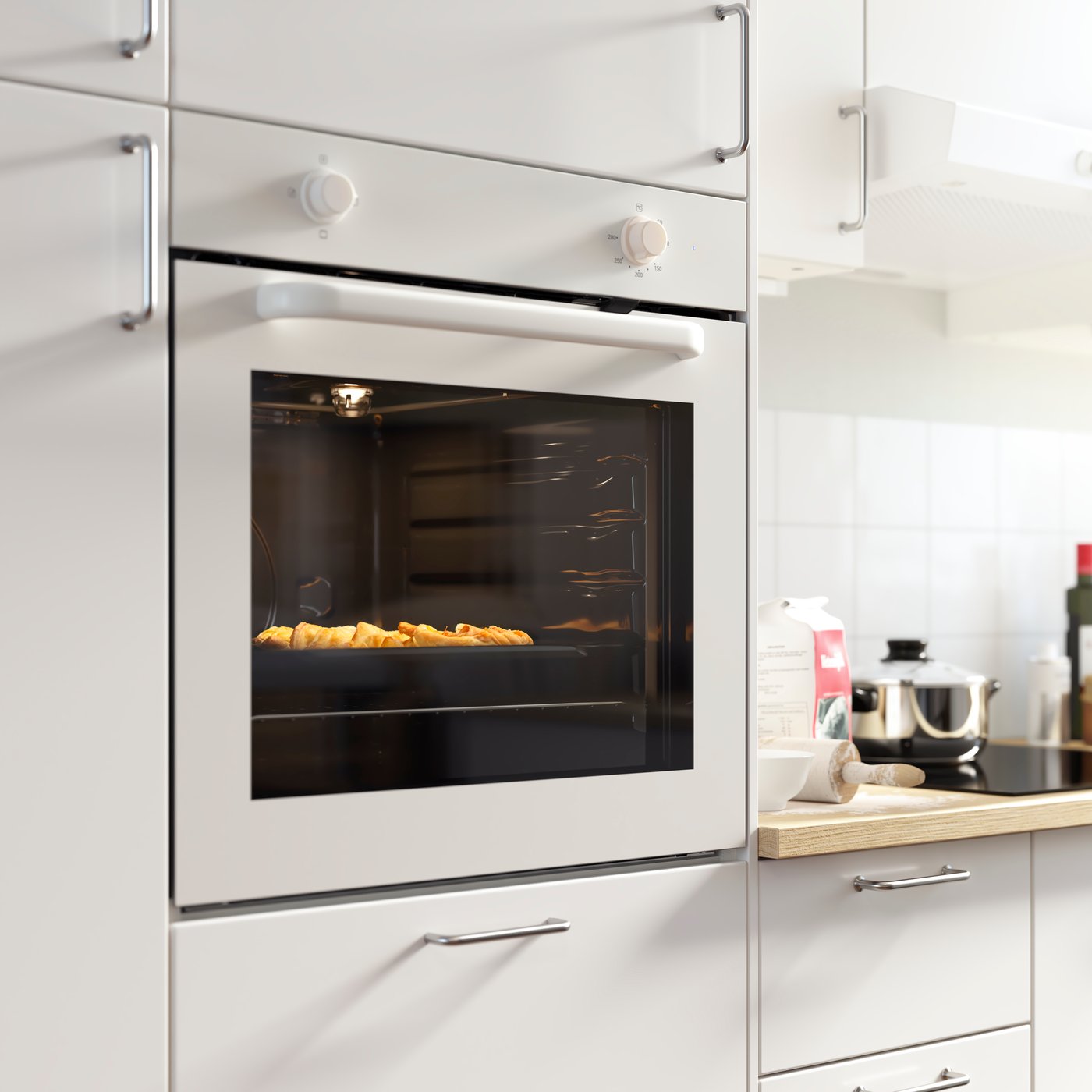 A white LAGAN oven is mounted in a bright kitchen with white kitchen fronts. The oven is turned on and there’s food baking.