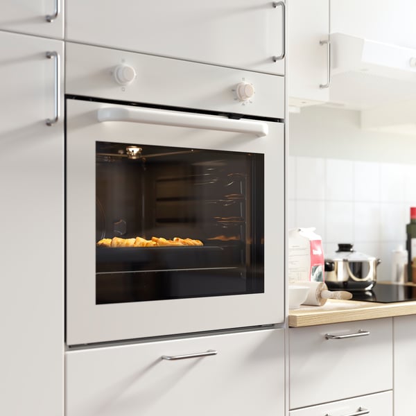 A white LAGAN oven is mounted in a bright kitchen with white kitchen fronts. The oven is turned on and there’s food baking.