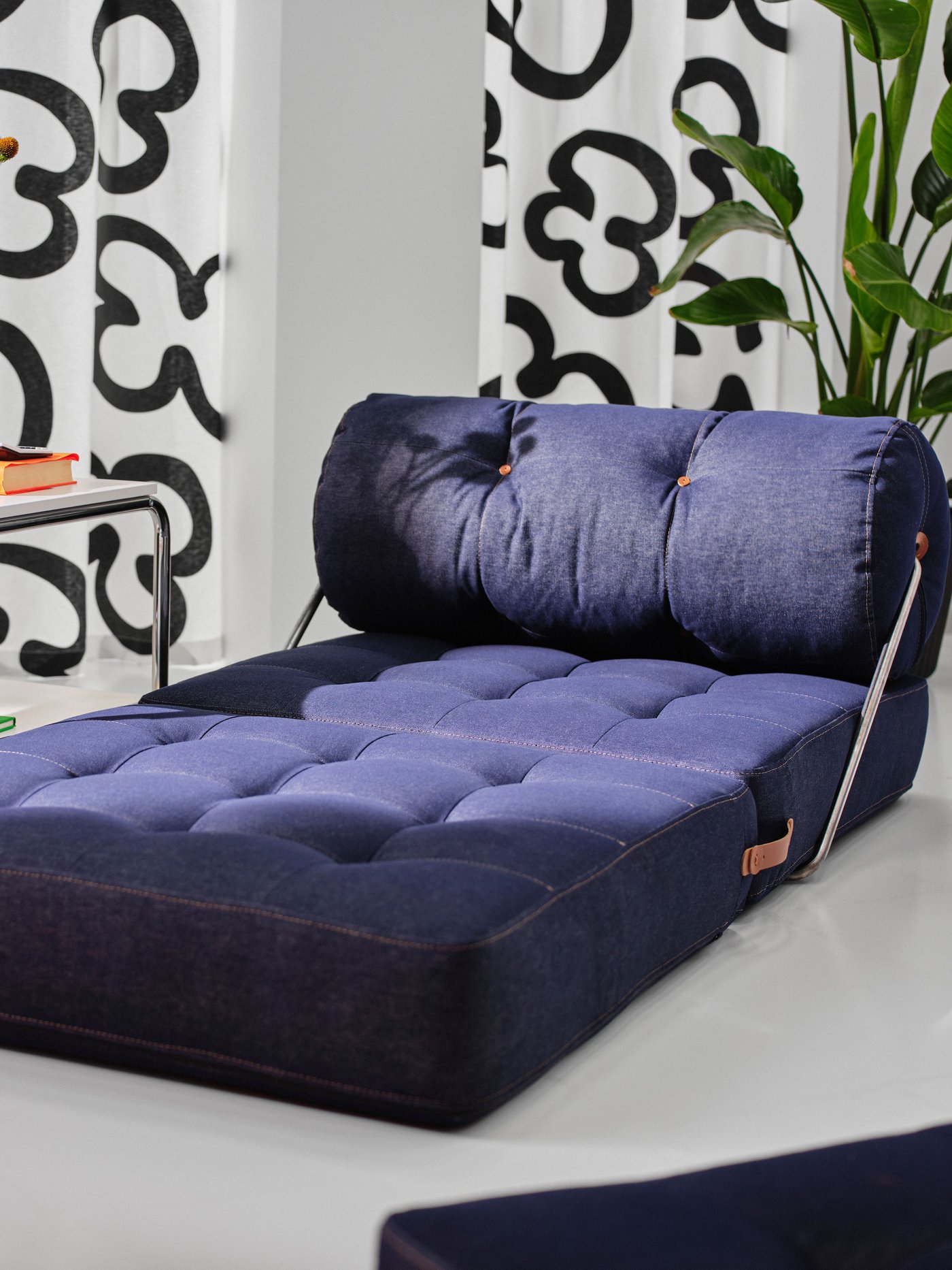 Mörkblå VÅRKUMLA fåtölj med tuftad sits och ryggstöd i ett ljust rum med svartvita gardiner och grön växt. Sidobord med bok och orange detalj syns bredvid.