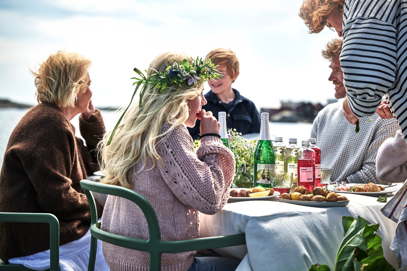 Une famille en train de manger en plein air, une petite fille avec une couronne de fleurs. Des boissons IKEA ISKUB et des boulettes de viande sur la table.