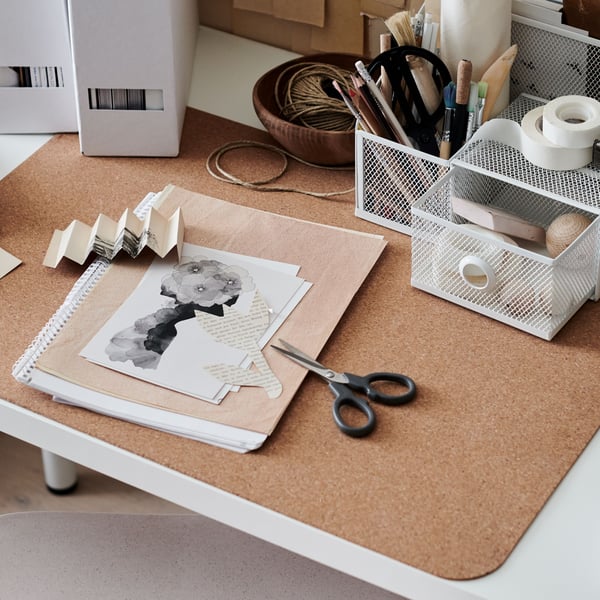 A notebook, scissors and a DRÖNJÖNS desk organiser on a cork SUSIG desk pad which is on top of a desk.