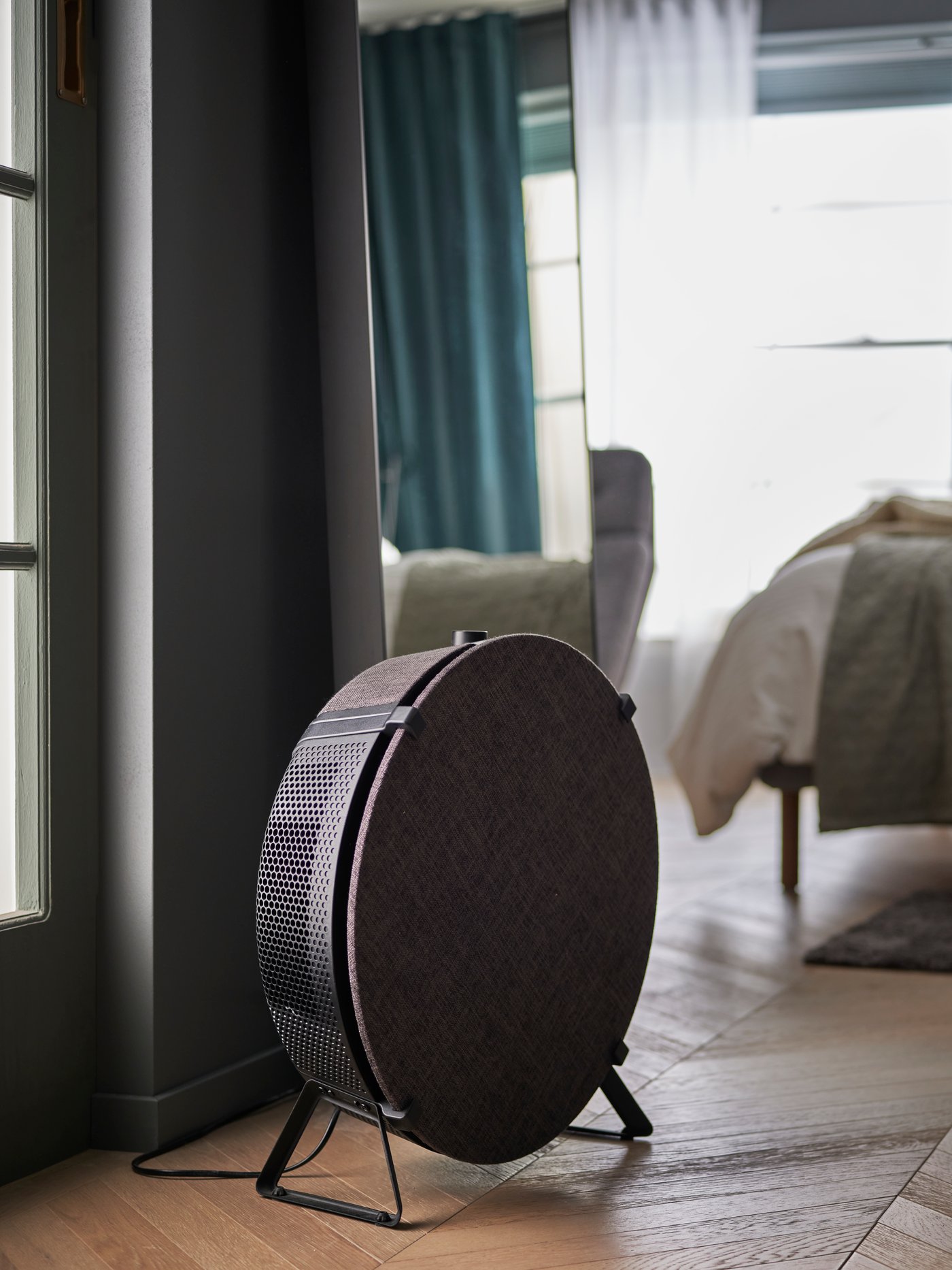 Close up on STARKVIND air purifier in black, standing on the floor with a mirror and a bed in the bedroom behind it.