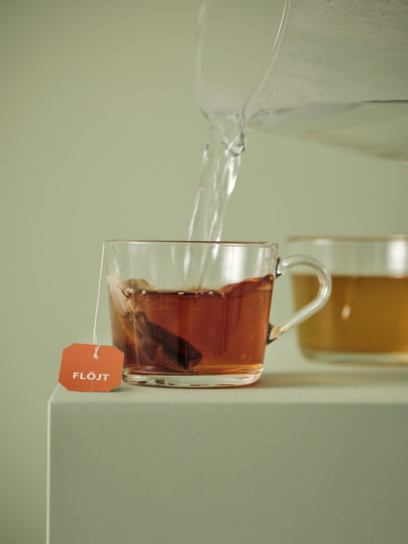 A small glass mug has a FLÖJT black teabag steeping inside it. Water is being poured from a large glass jug.