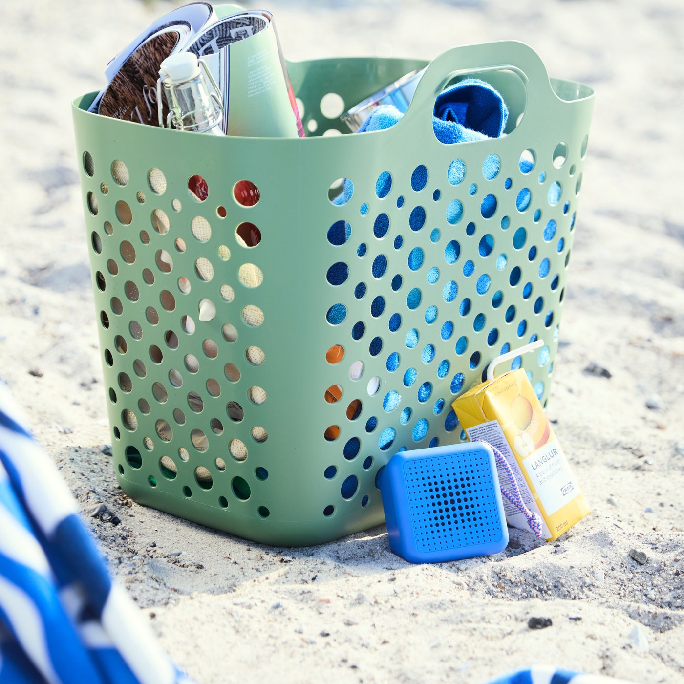En grön SLIBB flexibel tvättkorg fylld med strandsaker i sanden. Intill korgen lutar en blå VAPPEBY högtalare och ett smoothiepaket.