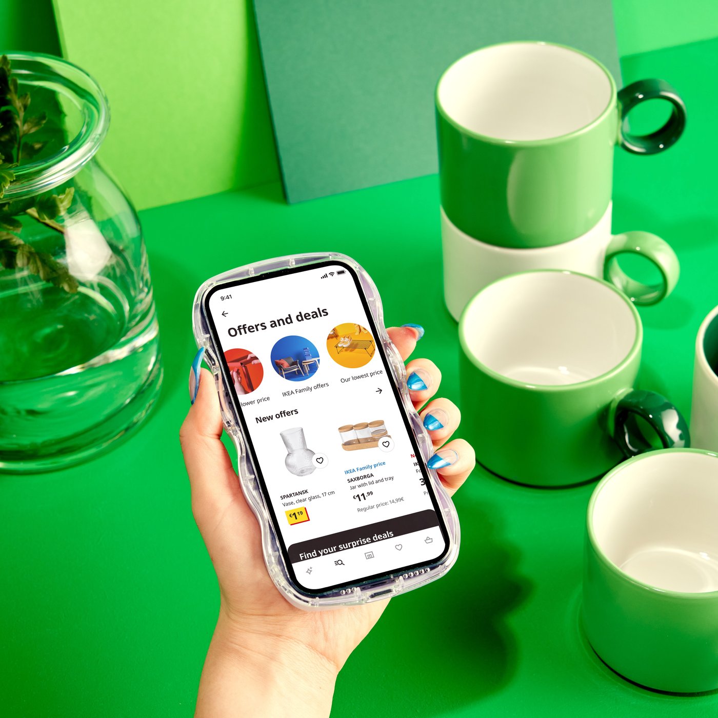 A hand is holding a smartphone displaying the IKEA app over a green table with green and white mugs.