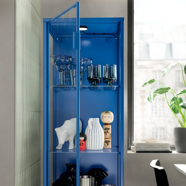 A blue RUDSTA glass cabinet next to a bright window in a living room.