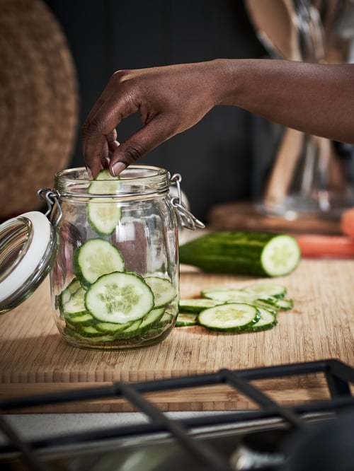 Kitchen with chopping boards, kitchen appliances, cooking utensils, jar with lid, hand placing sliced cucumber in the jar.