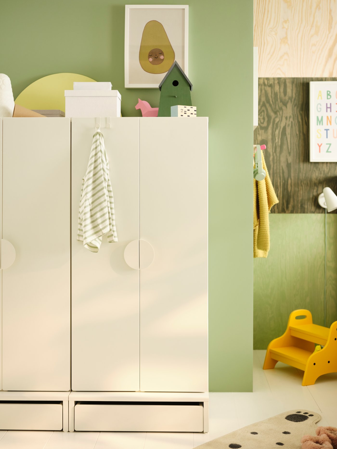 Armoire-penderie SMÅSTAD blanche installée contre un mur vert clair. Repose-pieds jaune dans un coin de la pièce.