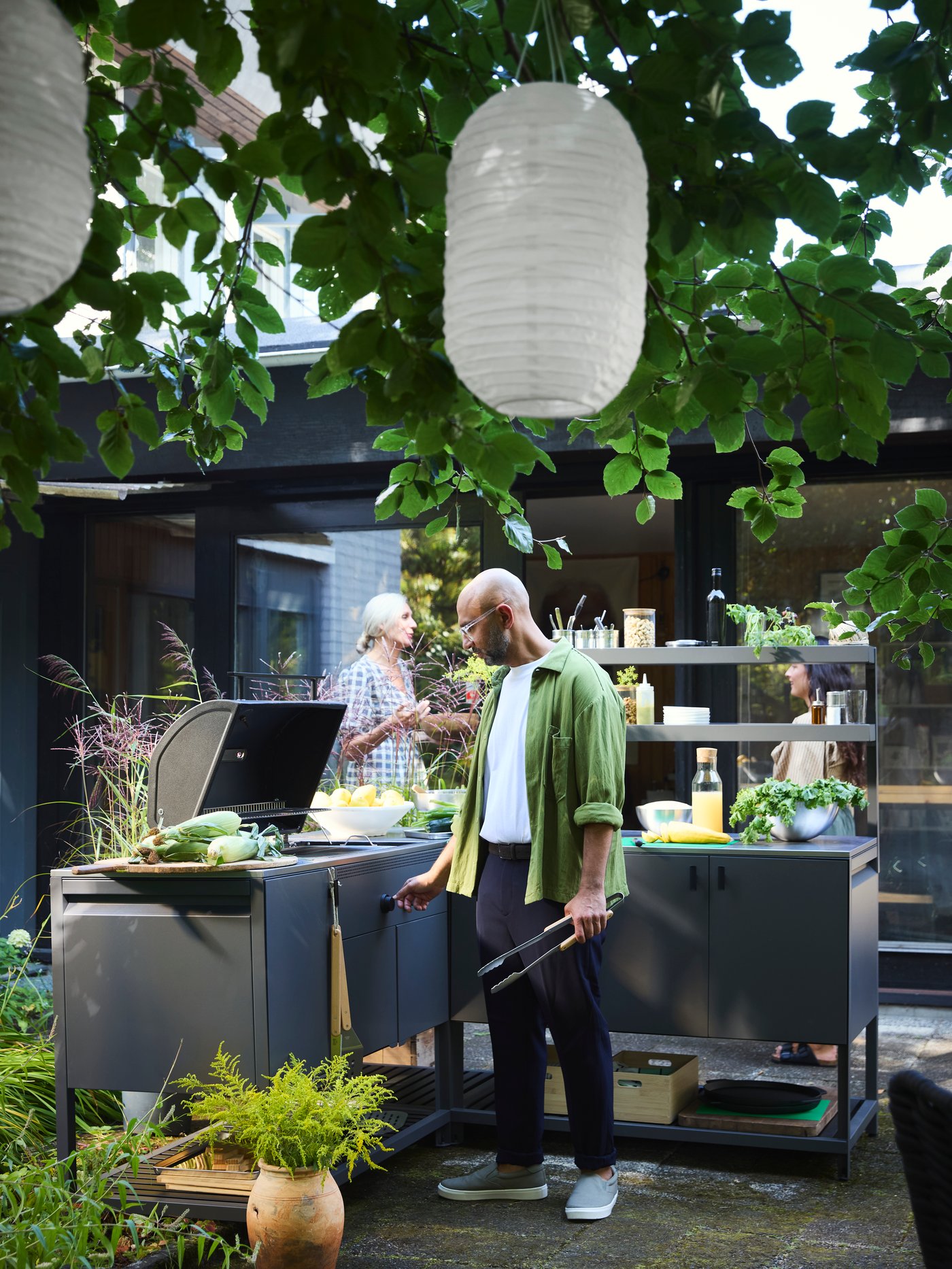 A man is in a garden and tends the barbecue in the BÅTSKÄR outdoor kitchen while two women chat in the background.