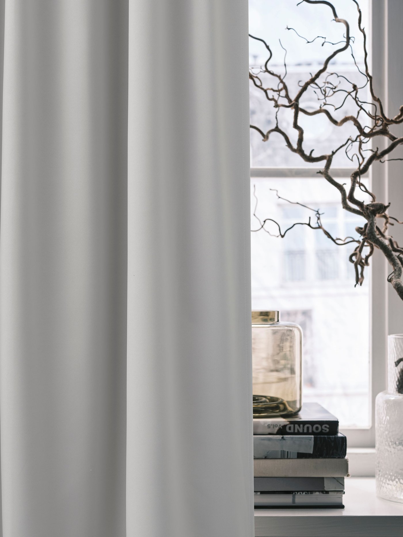 Ljusa rumsmörkande gardiner vid ett fönster, med en dekorativ gren i en vas bredvid.