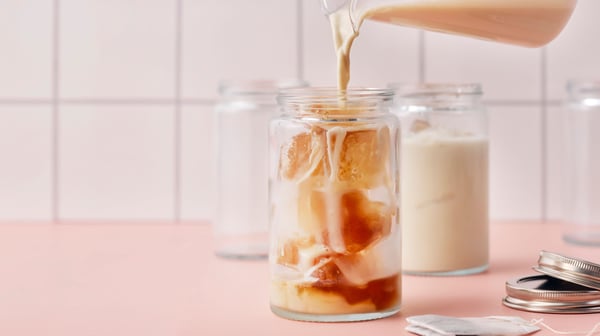Someone is pouring oat drink for coffee from a jug into a glass jar filled with ice cubes made of FLÖJT black tea and water.