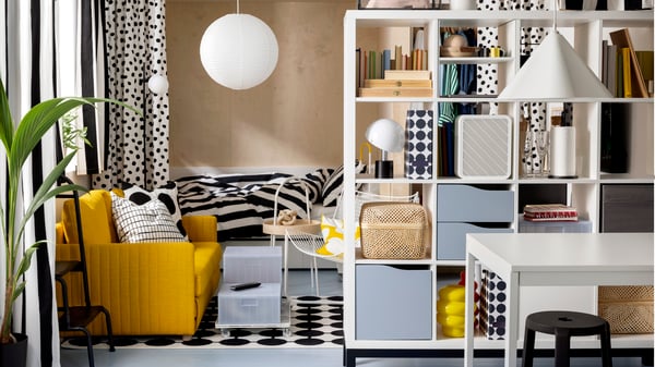 Convertible FRIDHULT dans un espace séjour près d’une étagère KALLAX blanche et d’un lit avec du linge de lit rayé.