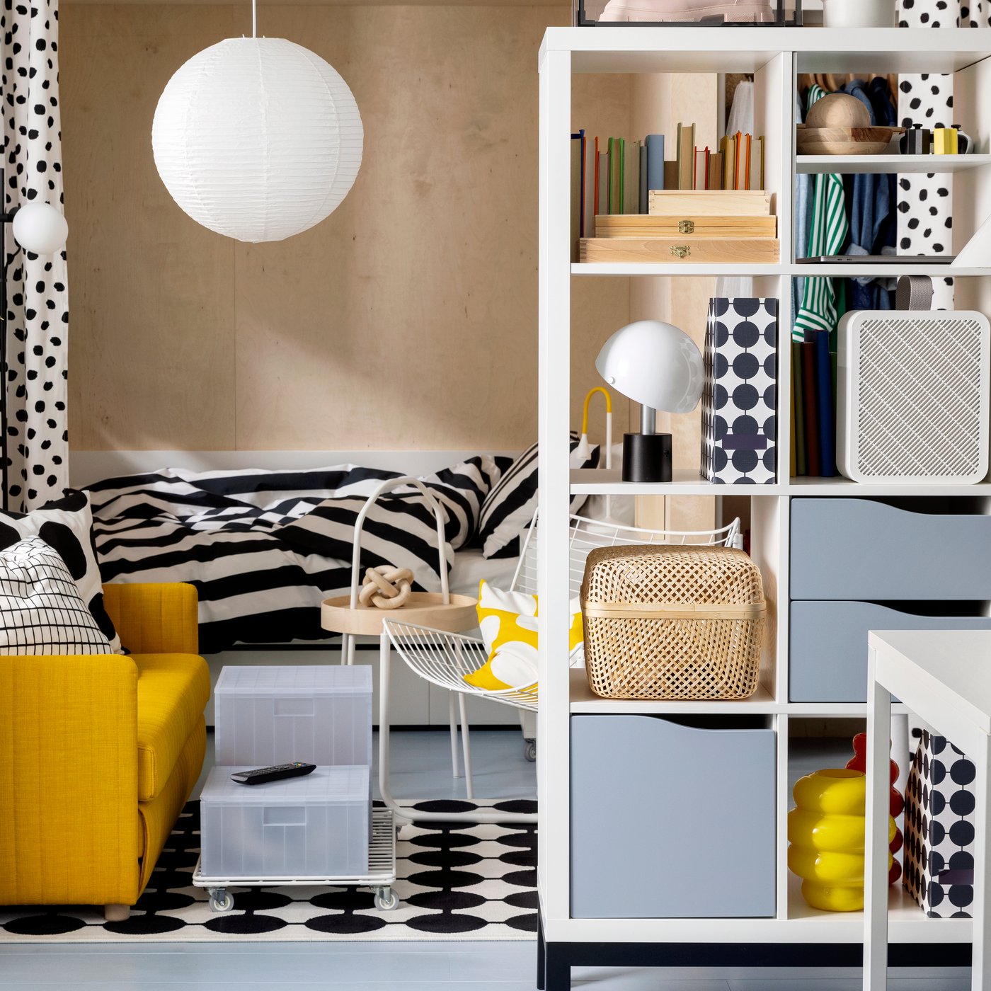 A FRIDHULT sofa-bed stands in a living room area near a white KALLAX shelving unit and a bed with striped bed linen.
