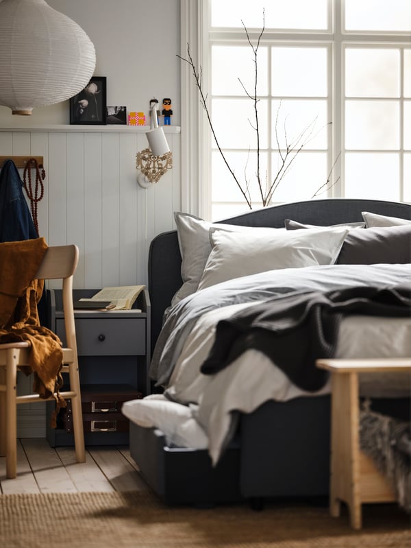 Una cama HAUGA con ropa de cama BLÅVINDA y una silla RÖNNINGE de abedul a su lado, delante de un gran ventanal.