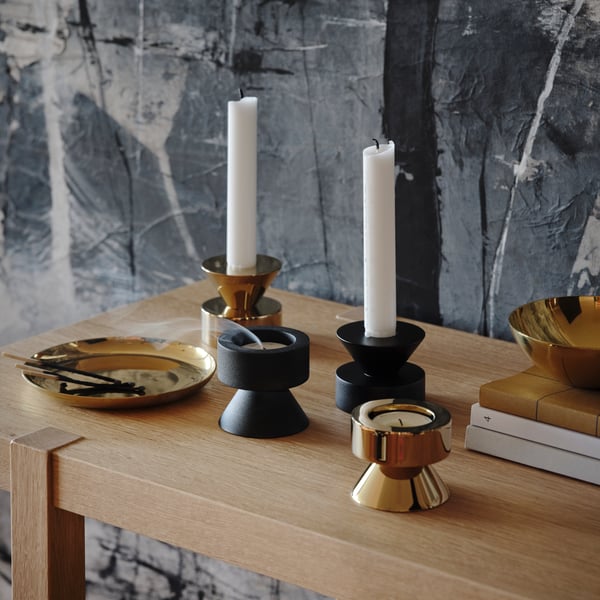 Brass-coloured and black STOCKHOLM 2025 candlestick/tealight holders with candles and tealights on an oak surface.