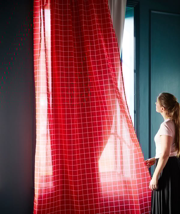 A woman holds red curtains and looks out as the sunlight comes in through the window.