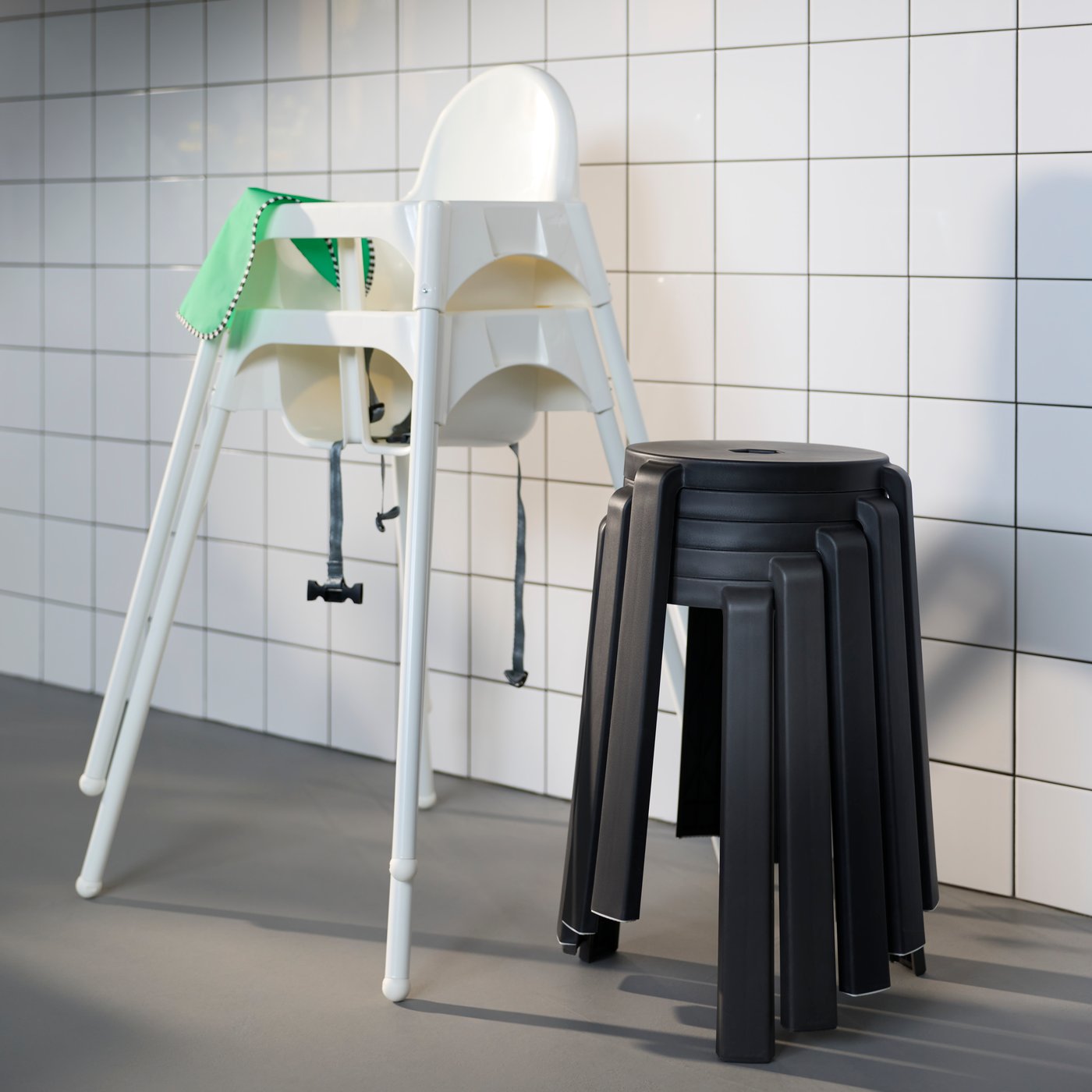 Dvě bílé vysoké židle ANTILOP s bezpečnostními pásy stojí u obložené stěny vedle sady černých stoliček.