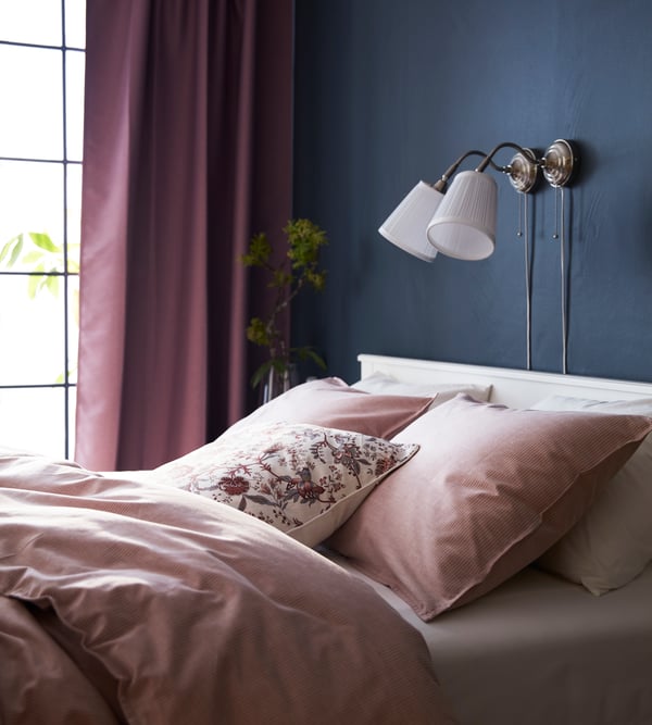 Zwei ÅRSTID Wandleuchten mit silbernen Sockel hängen über einem romantisch, traditionellen Bett in weiß und rosé.