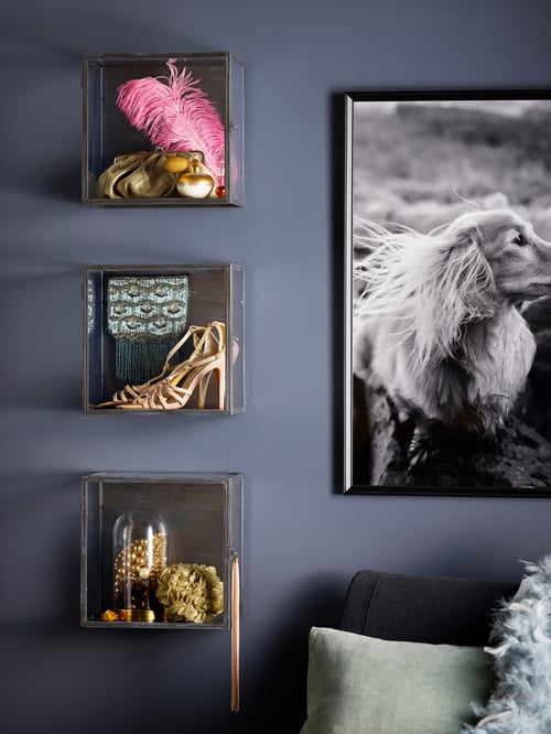 Trois boîtes vitrées BARKHYTTAN sont fixées à un mur gris foncé. Elles contiennent des accessoires dorés, des talons aiguilles et une plume rose.