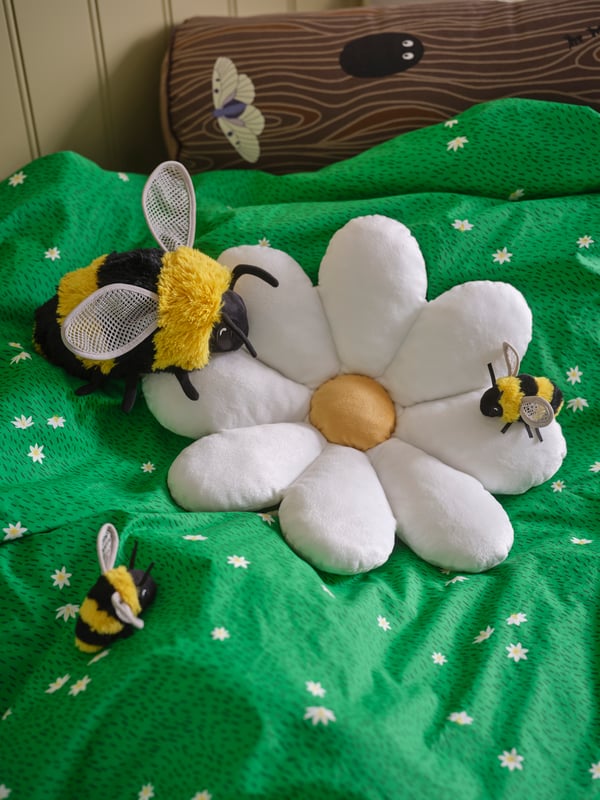 A SKOGSDUVA flower-shaped cushion, one SKOGSDUVA bumblebee soft toy and two mini-bumblebee soft toys are placed on a bed.