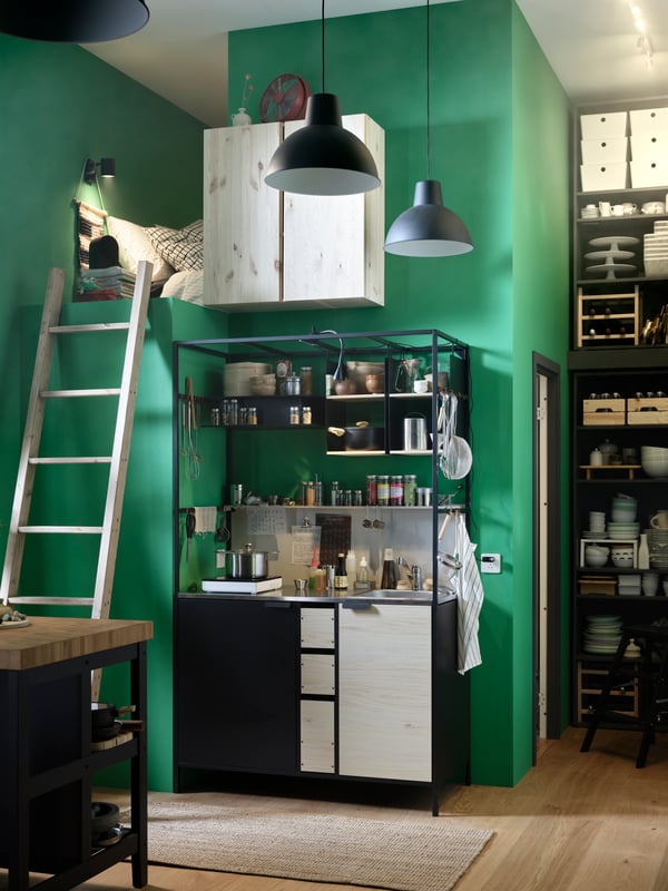 An ÄSPINGE kitchenette with utensils, spices, and more sits against a green wall next to a ladder leading to a sleeping loft.
