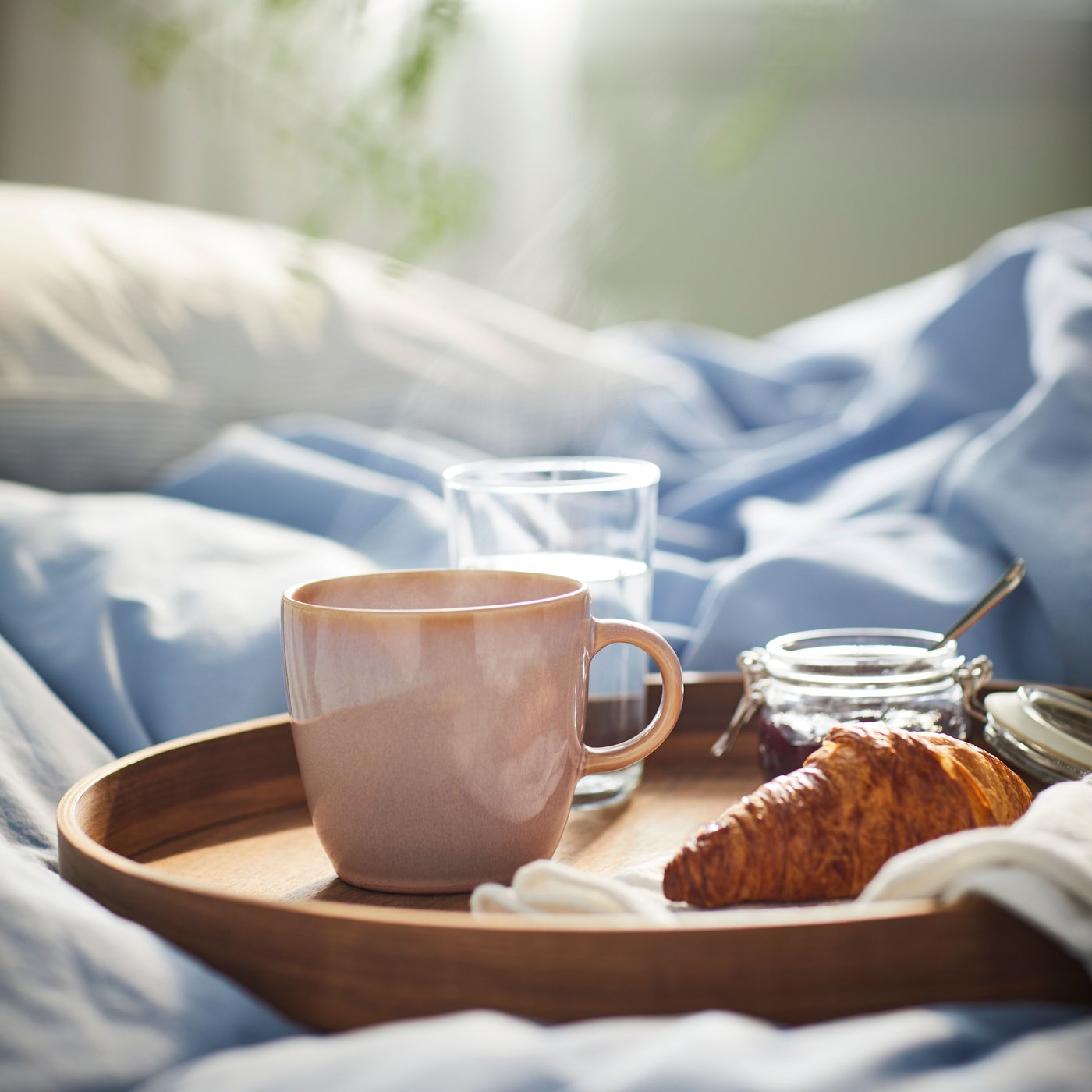 Holztablett mit einer hellrosanen FÄRGKLAR Tasse, Wasserglas, Marmeladenglas und Croissant auf einem Bett mit blauer Bettwäsche – ein gemütliches Frühstück im Bett im Morgenlicht.
