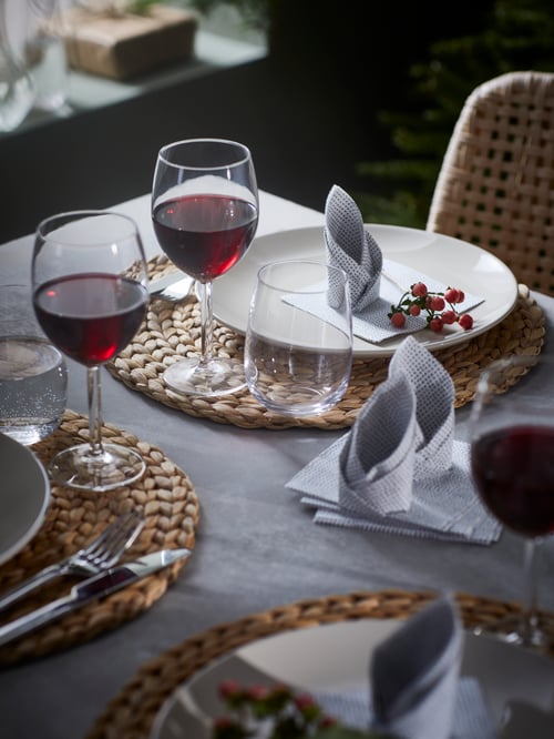 A decorated table set with braided SOARÉ place mats, folded napkins, beige FÄRGKLAR plates and drinks in SVALKA wine glasses.