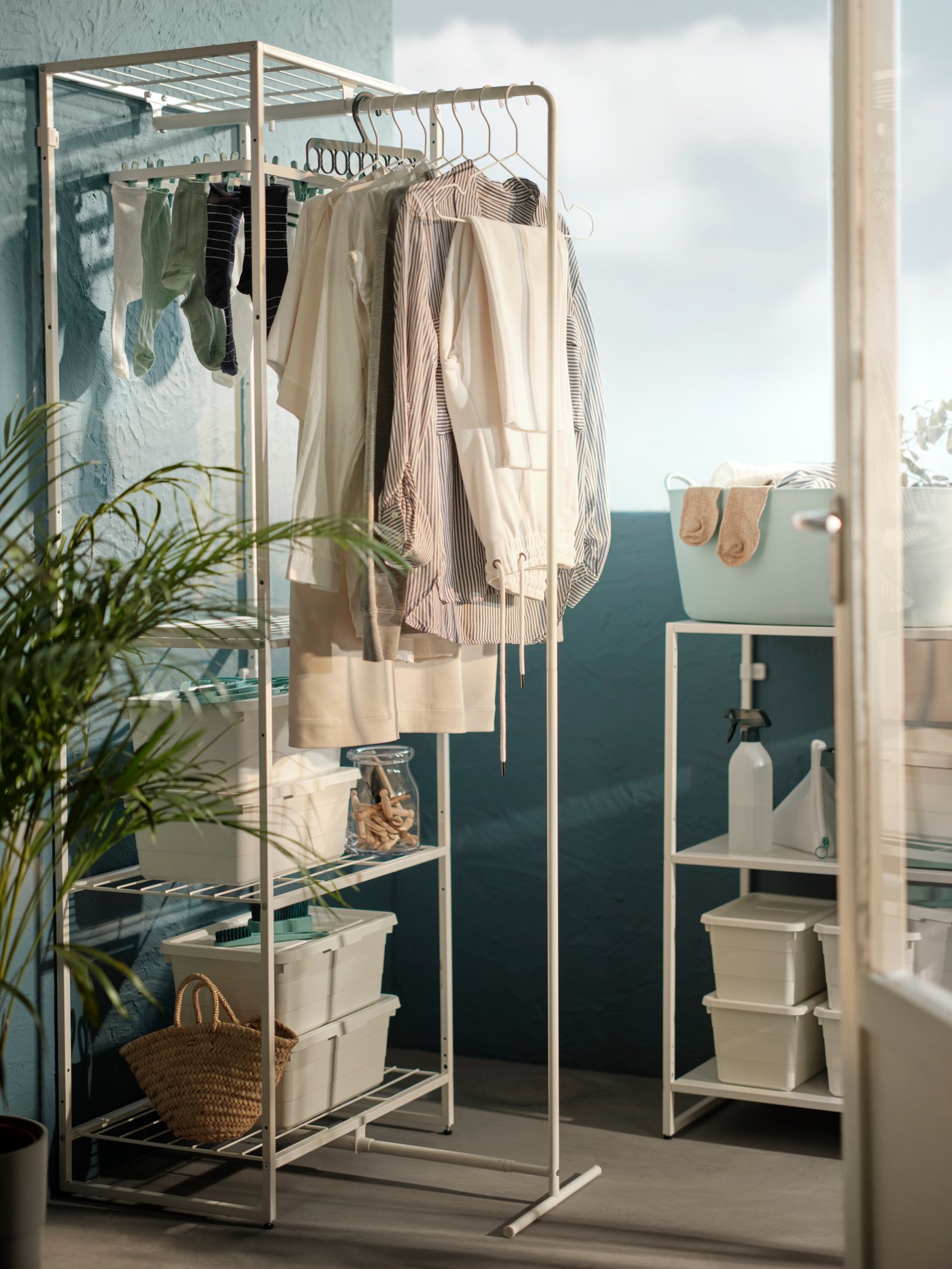An outdoor laundry storage setting featuring a JOSTEIN shelving unit with a drying rack placed against a blue wall and another JOSTEIN shelving unit with a TORKIS laundry basket.