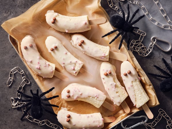 Frosty banana ghost popsicles dipped in BELÖNING white chocolate with raspberries, with toy spiders and chains around them.