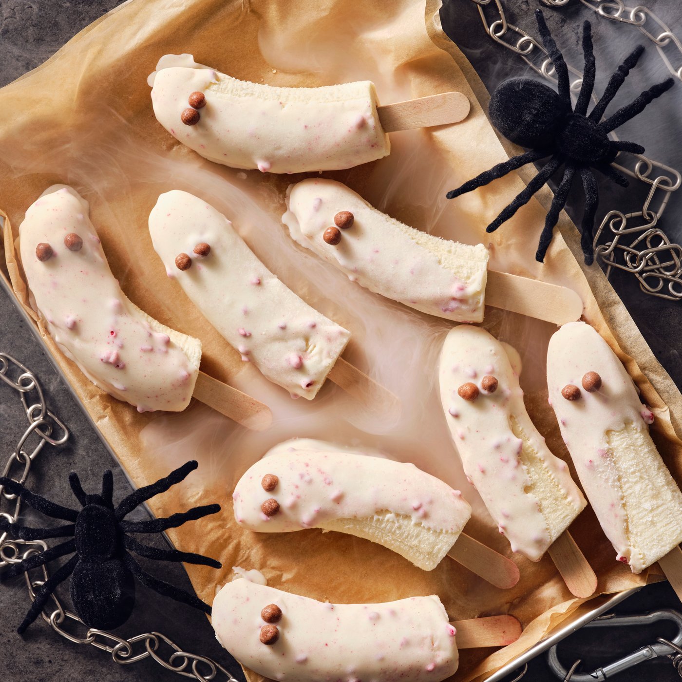 Frosty banana ghost popsicles dipped in BELÖNING white chocolate with raspberries, with toy spiders and chains around them.