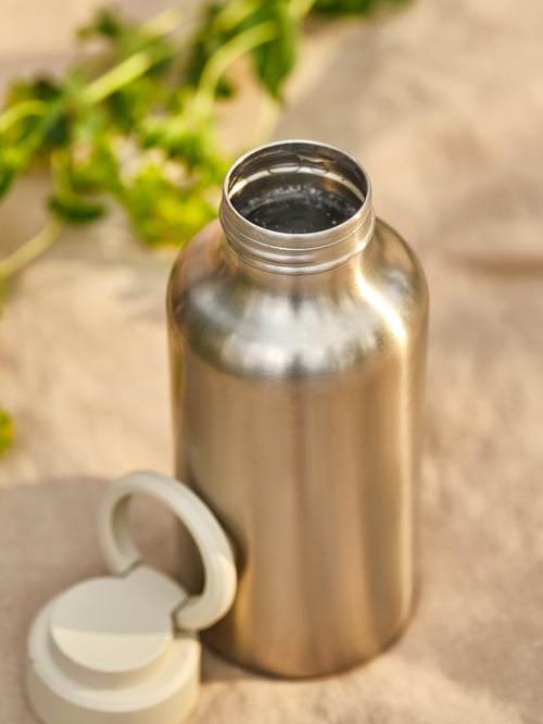 Eine ENKELSPÅRIG Edelstahlwasserflasche mit beigefarbenem Verschluss steht auf einer sonnenbeschienenen Oberfläche.