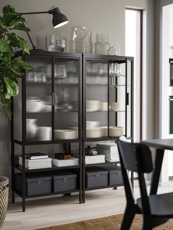 Two MOSSJÖN glass-door cabinets in anthracite have plates and glasses inside, vases on top and boxes on the lower shelves.