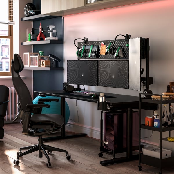 A black MÅLOMRÅDE sit/stand gaming desk is featured in a stylish room with shelves, a gaming chair and gaming gear.