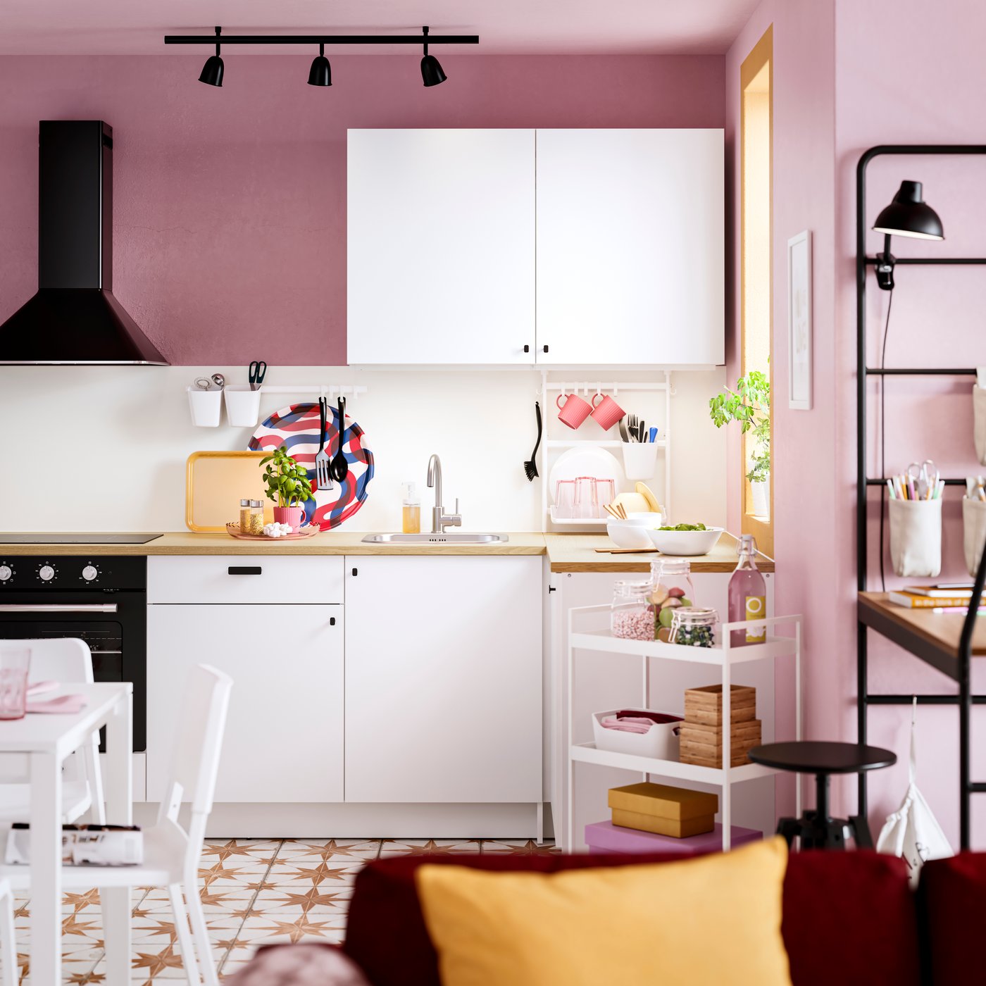 Kompakte IKEA KNOXHULT Küche mit weißen Fronten und Holzarbeitsplatte in einem rosa gestrichenen Raum, schwarzer Dunstabzugshaube und Schienenspot, ergänzt durch Servierwagen, Esstisch und farbenfrohe Accessoires für ein fröhliches, kreatives Wohngefühl.