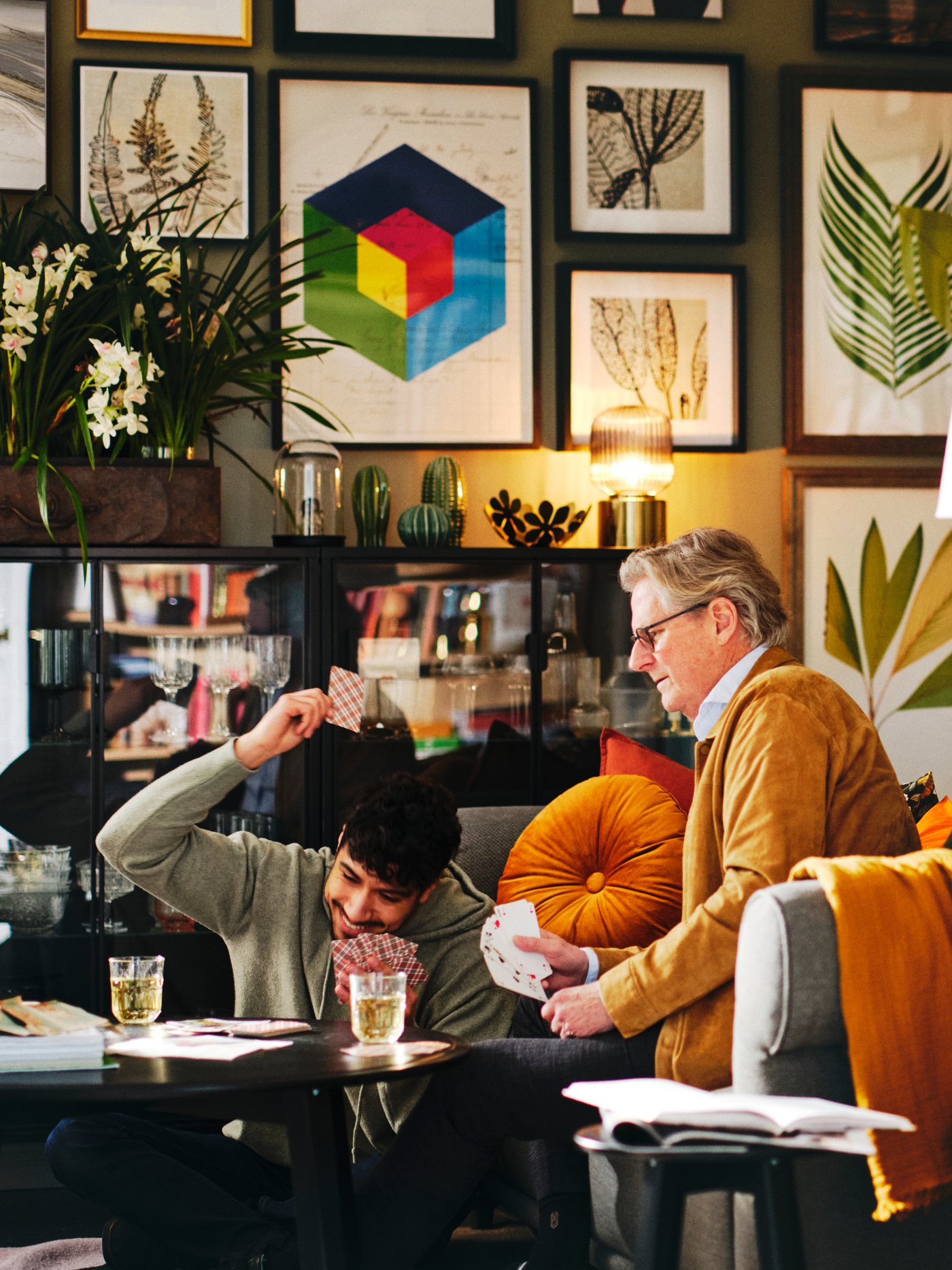 Młody mężczyzna grający w karty ze starszym mężczyzną siedzącym na sofie w pokoju dziennym z galerią obrazów na ścianie.