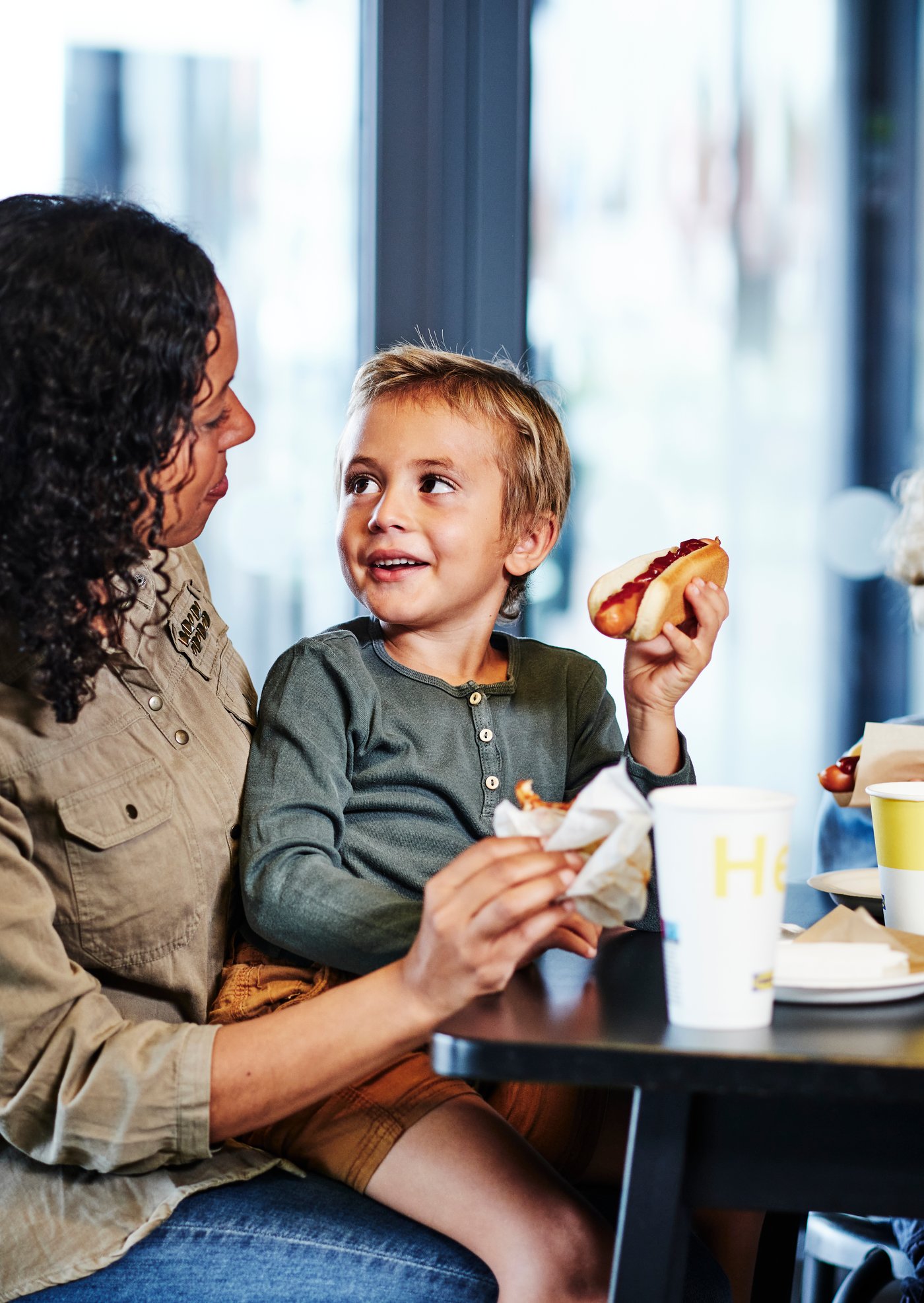 A mother and child sit at the table, with the child on the mother’s lap, eating a hot dog.